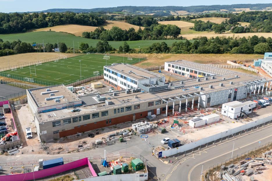 Aerial view of Matford Brook Academy, an unused school in Exeter, showing the building under construction and a sports field nearby, with green hills in the background.