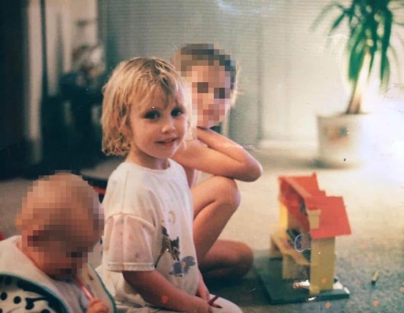 A young child with blonde hair smiles at the camera, with a partially pixelated older child behind them, and a partially pixelated baby to their left, all seated on a floor with a toy house.