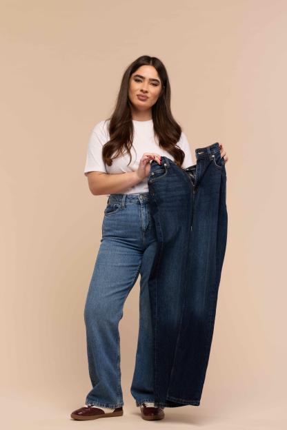 A woman models light blue jeans, holding a pair of dark blue jeans.
