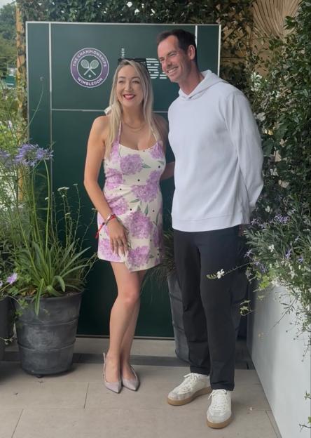 Andy Murray poses with a woman in a floral dress in front of a Wimbledon sign.