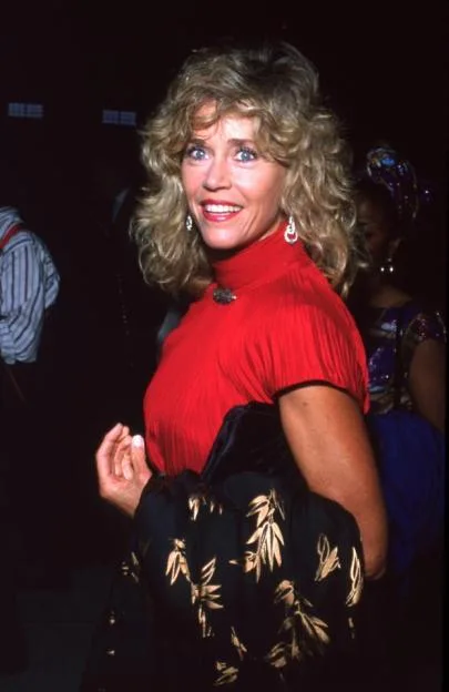 Jane Fonda smiling at the camera, wearing a red top and holding a black garment with gold leaf patterns.