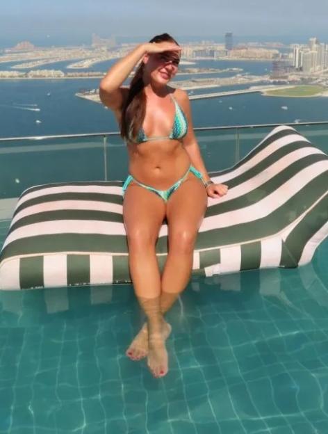 A woman in a bikini sits on a lounge chair in a rooftop pool overlooking a city and coastline.