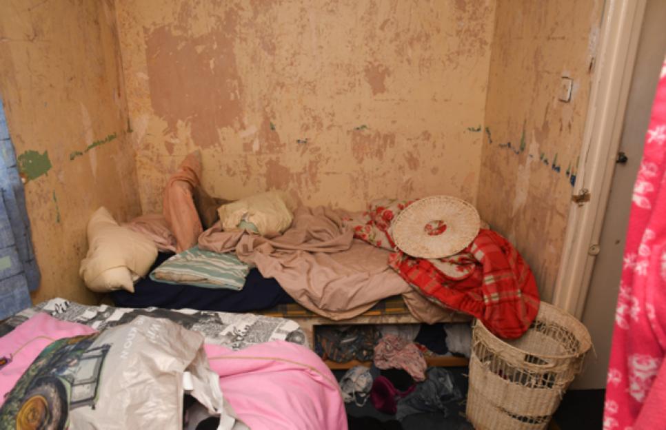 A small, squalid bedroom with peeling wallpaper, a bed piled with blankets and pillows, and a wicker laundry basket next to it.