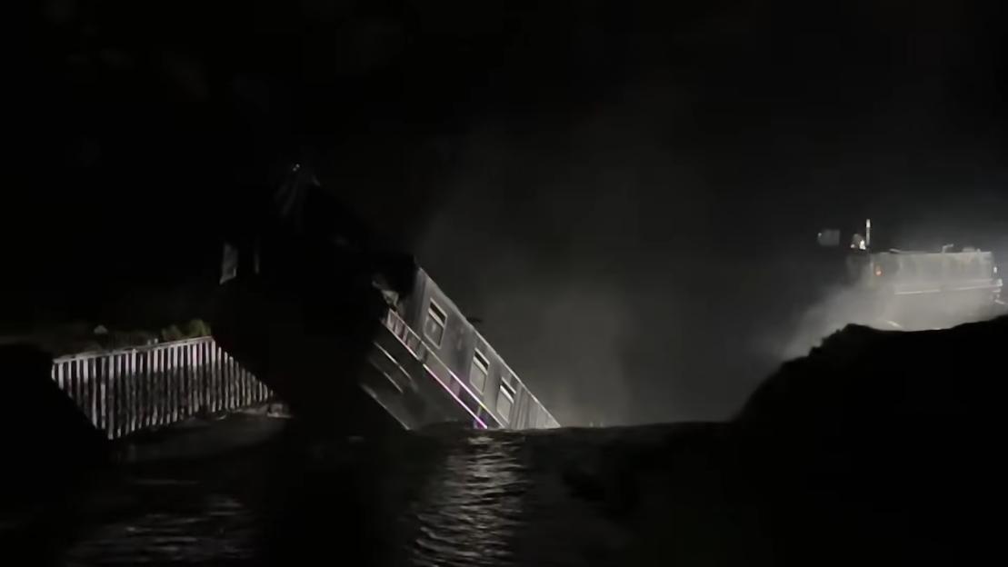 A canal boat partially submerged at a steep angle in a sinkhole at night, with a rescue vehicle in the background.