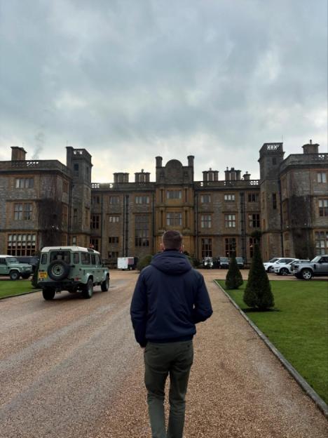 A person walks up a gravel driveway toward a large, historic manor house with several cars parked on either side.