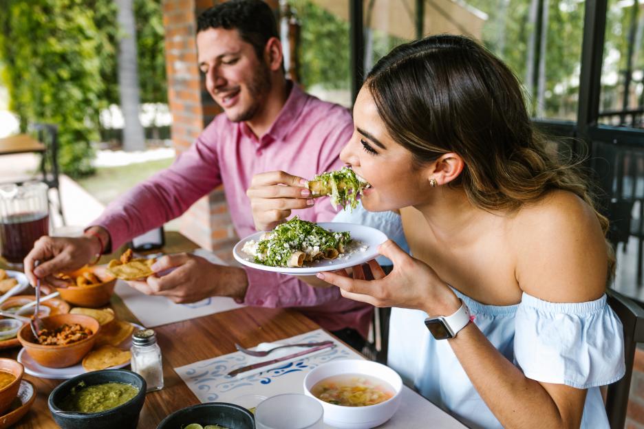 A young Latin woman eating Mexican tacos on a restaurant terrace, with a young man in the background.