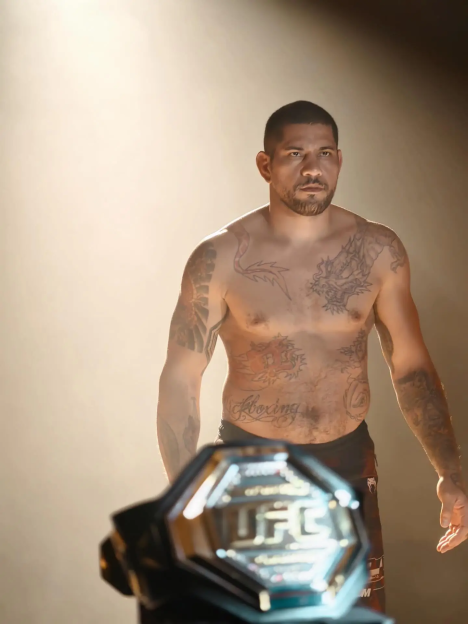 A male fighter with numerous tattoos, shirtless, standing under a spotlight with a championship belt in the foreground.