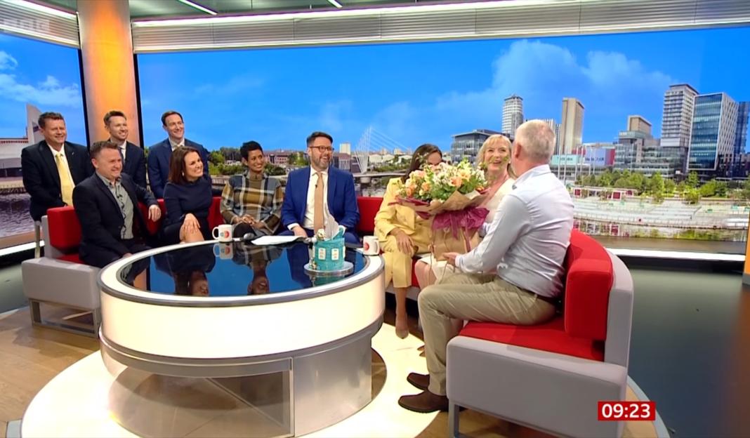 A group of people sitting on a TV set with a city skyline in the background, one woman holding a bouquet of flowers and a man offering her a gift.