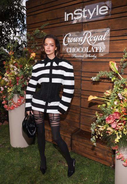 Maura Higgins in a black and white striped dress with a black corset over it, black patterned tights, and black heels, holding a black handbag, standing in front of a wooden wall with "InStyle" and "Crown Royal Chocolate" logos.