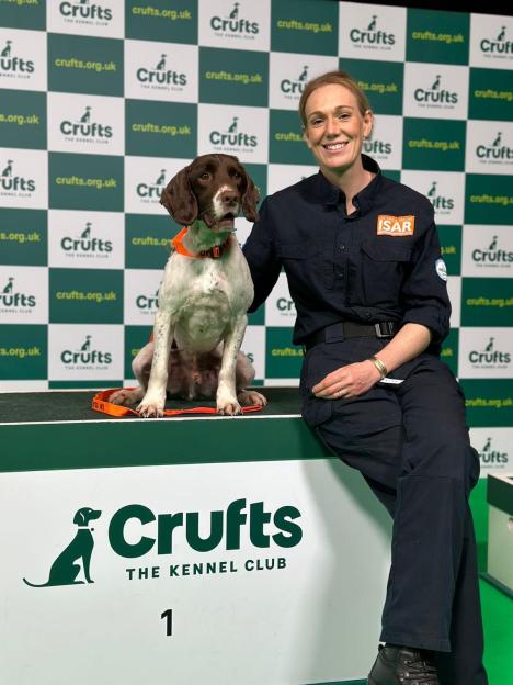 A woman in a dark uniform with "UK FIRE & RESCUE ISAR" patches sits next to a brown and white dog on a podium with "Crufts The Kennel Club 1" written on it.