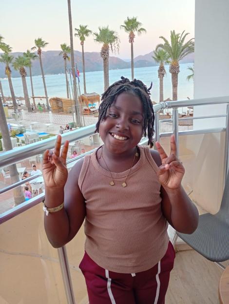 Eva, 9, smiling and holding up two peace signs, on a balcony overlooking a beach with palm trees and water.