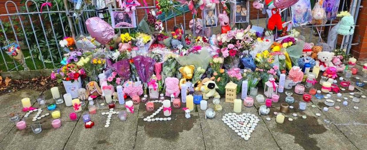 A memorial with flowers, candles, teddy bears, and a pink balloon on a sidewalk next to a fence.