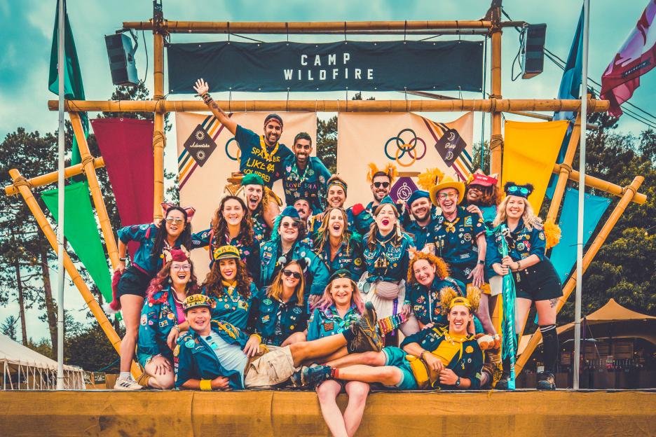 A large group of smiling adults in camp uniforms with badges and colorful accessories pose for a photo under a “Camp Wildfire” banner.