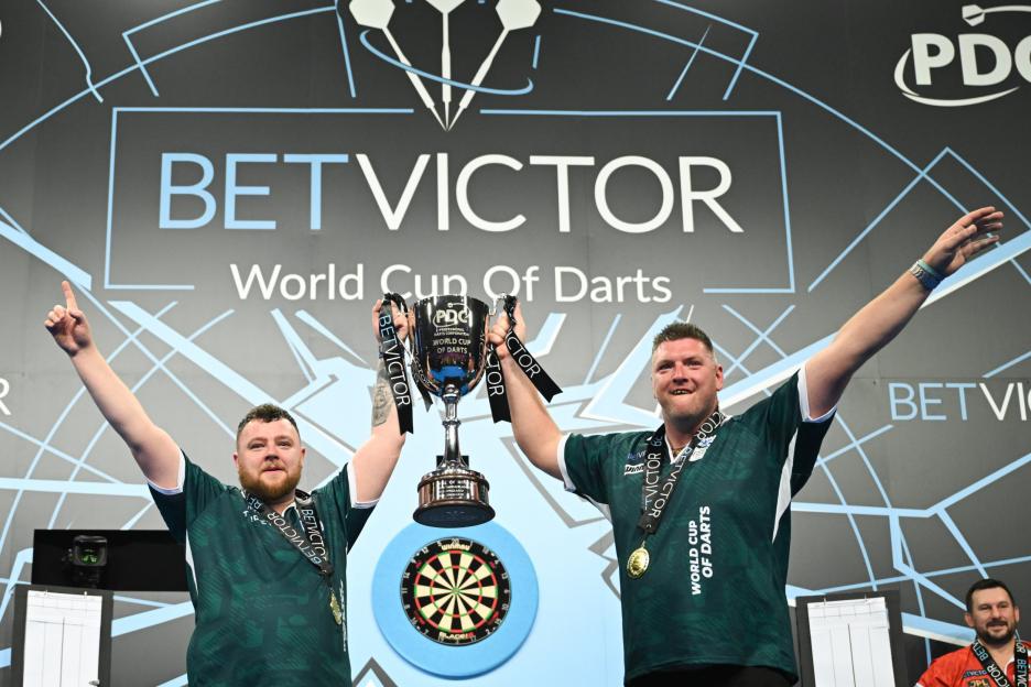 Two male dart players raise the BetVictor World Cup of Darts trophy.