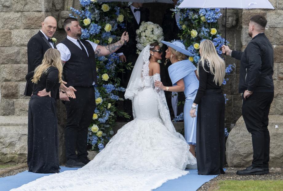 A bride and another woman in a blue dress and hat touching noses, surrounded by several people outside a venue decorated with blue and yellow flowers.