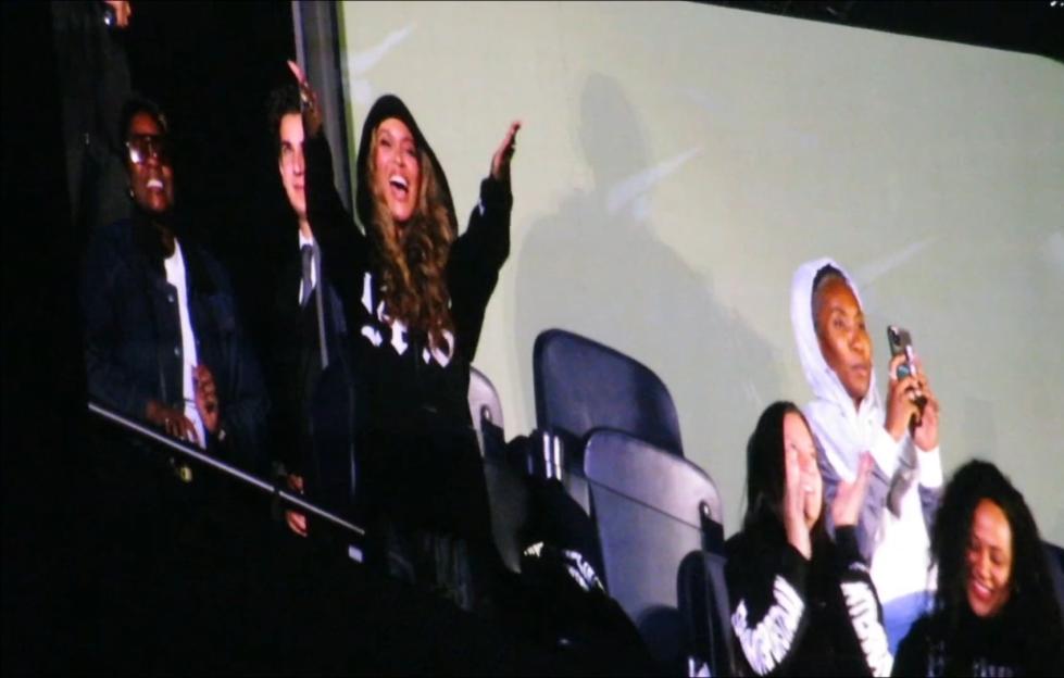Beyoncé smiles with her arms raised while Jay-Z and others in the crowd watch her.
