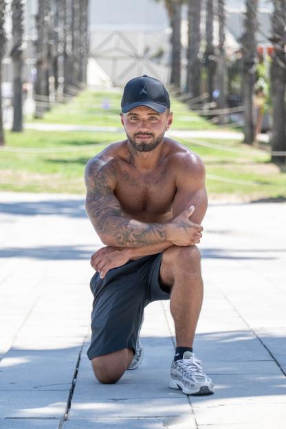 Tommo Holbrook, shirtless and tattooed, kneeling outdoors.