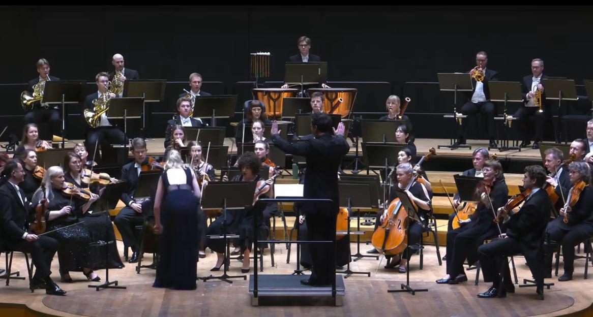 Conductor Matthew Halls on a podium directs an orchestra, while violinist Elina Vähälä (left, wearing a long navy dress) walks towards him.