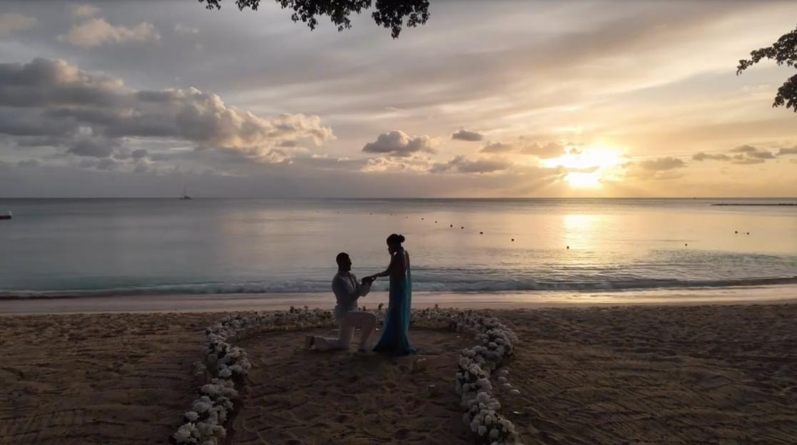 Man proposing to a woman on a beach at sunset, with white roses forming a heart on the sand.