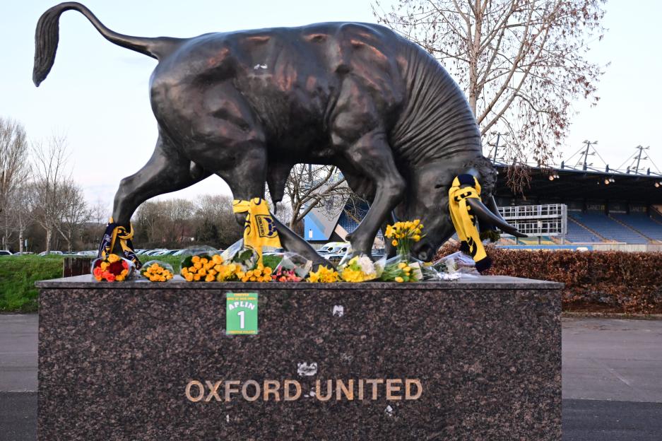 A bull statue adorned with yellow scarves and surrounded by bouquets of yellow and orange flowers, with an Oxford United sign and an Aplin 1 poster on its base.