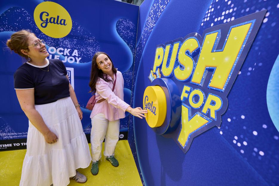 Two women interact with an oversized "Push for Joy" button and "Gala" branding at an event.