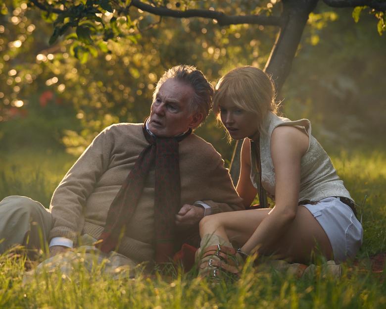 An older man and younger woman sitting in a grassy field with sunlight filtering through trees.