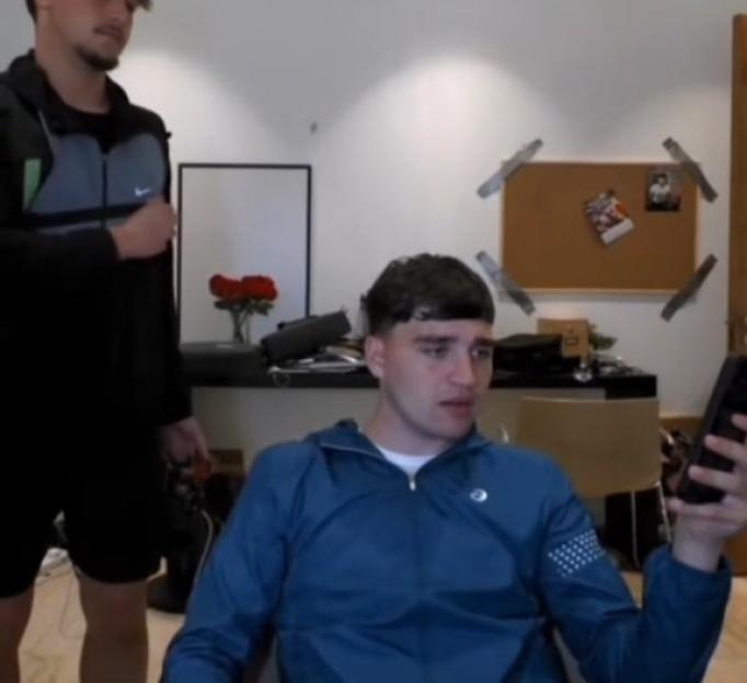 A man in a blue jacket looking at his phone, while another man stands behind him.