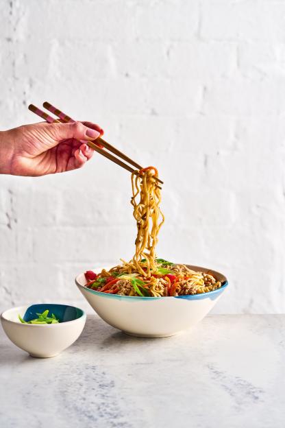 A hand holding pink and brown chopsticks lifts noodles from a large bowl of stir-fry, with a small bowl of green onions to the side.