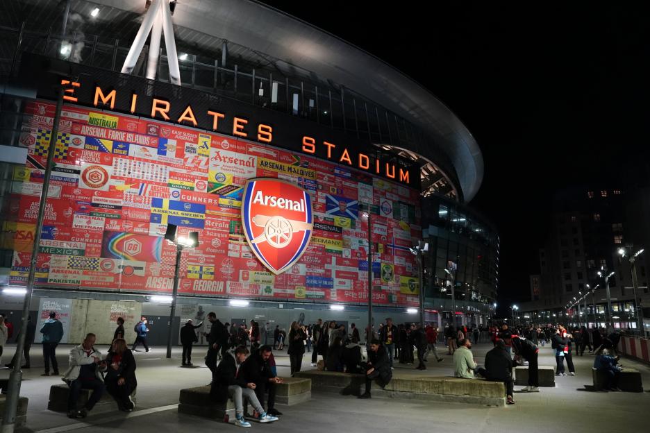 The Emirates Stadium at night, with a large Arsenal crest and various Arsenal supporters' club flags from around the world displayed on its exterior, and people gathered outside.