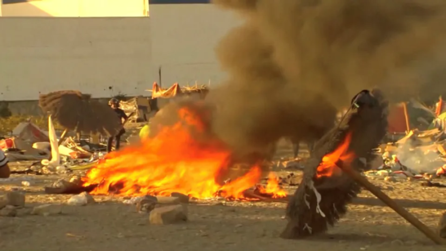 A large bonfire with thick smoke in a barren area, with debris and two people in the background.