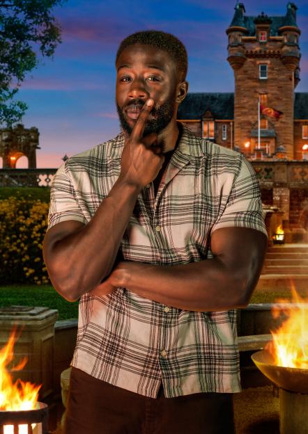 Ross, a Sales Exec and Personal Trainer from London, in a plaid shirt with his finger to his lips, with a castle and fire pits in the background.