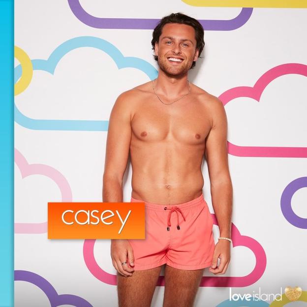 Casey from Love Island, in coral swim shorts, smiling in front of a cloud-patterned background.