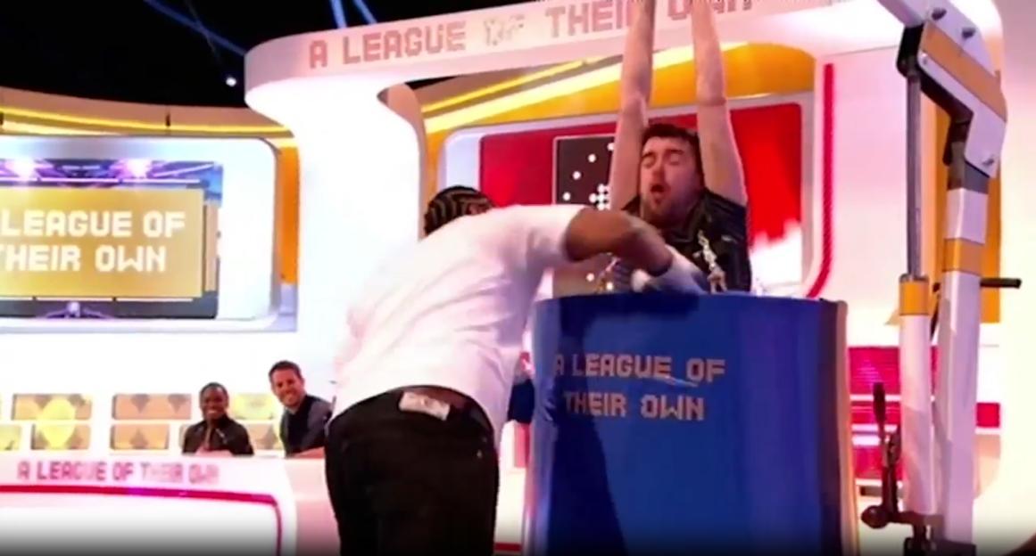 A man with his arms raised, standing in a blue barrel that says "A LEAGUE OF THEIR OWN," as another man leans over to interact with him in a game show setting.