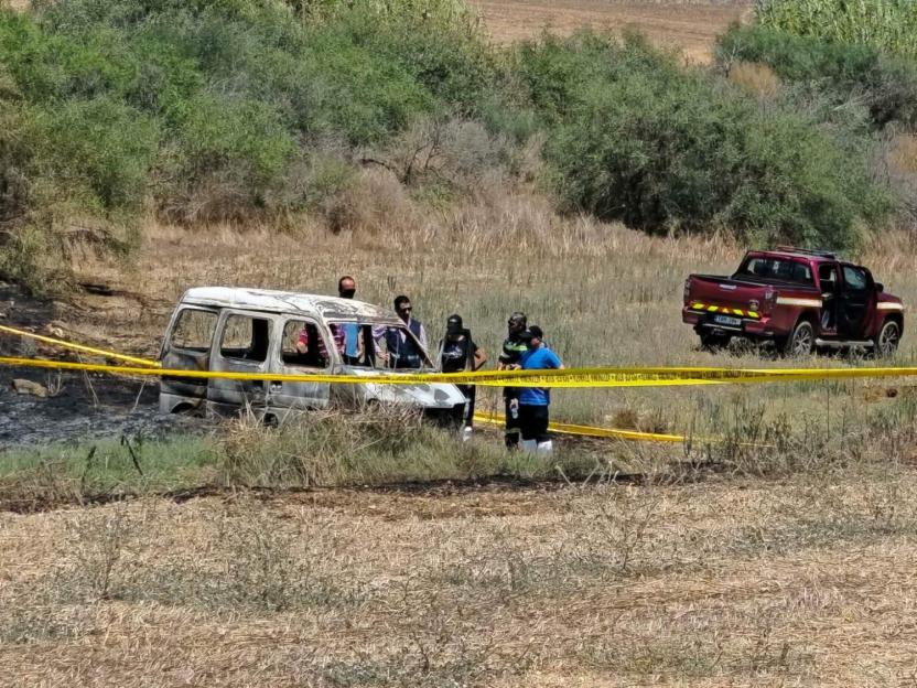 A burnt-out van surrounded by yellow police tape in a field with several individuals standing nearby, and a red pickup truck in the background.