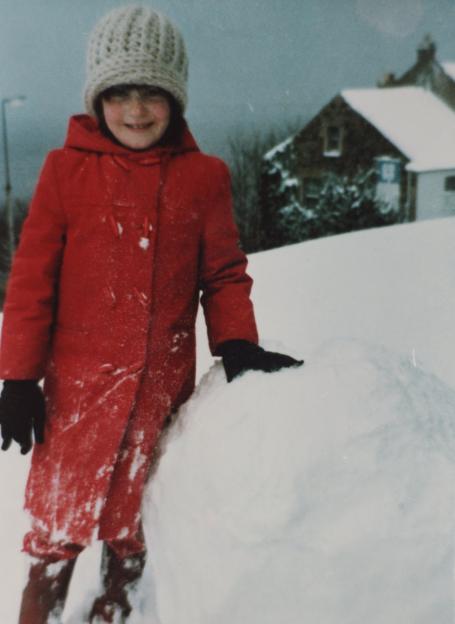 Susan Maxwell stands next to a large snowball in a snowy outdoor scene.