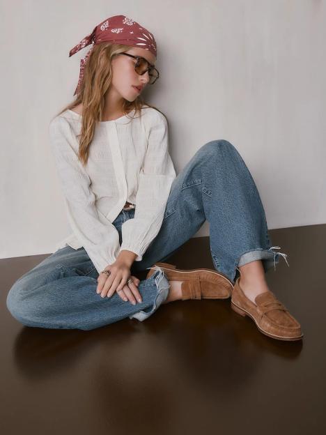 A woman in a white blouse, blue jeans, and brown loafers sits on a brown floor.