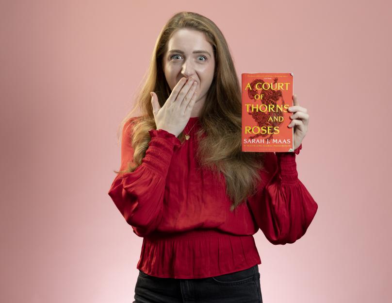 Emily Webber holding a copy of "A Court of Thorns and Roses" by Sarah J. Maas, looking surprised with her hand covering her mouth.