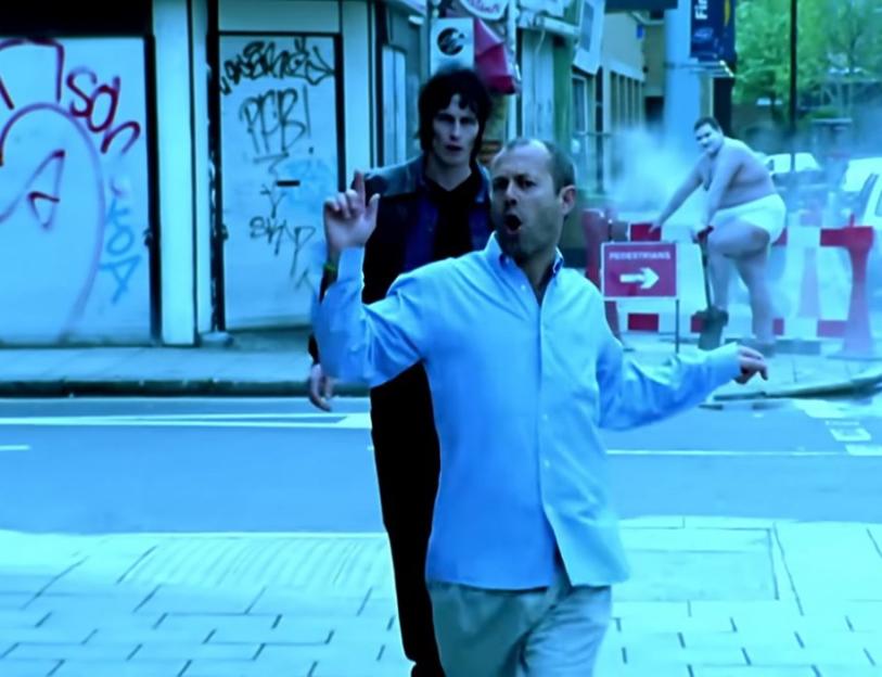 A man in a light blue shirt dancing in the street, with another man and a large person in a diaper behind him.