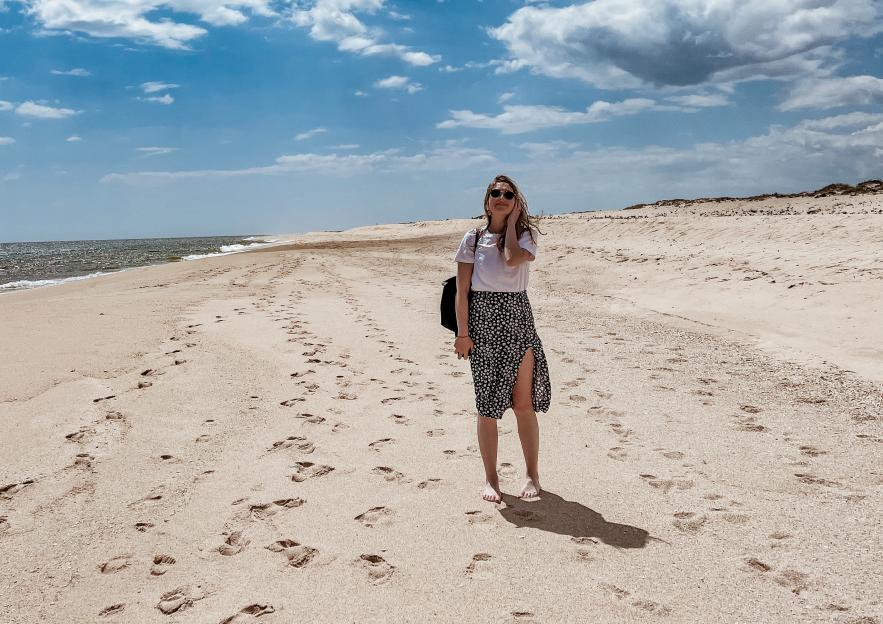 A person in sunglasses, a white shirt, and a long black floral skirt stands on a sandy beach next to the ocean.