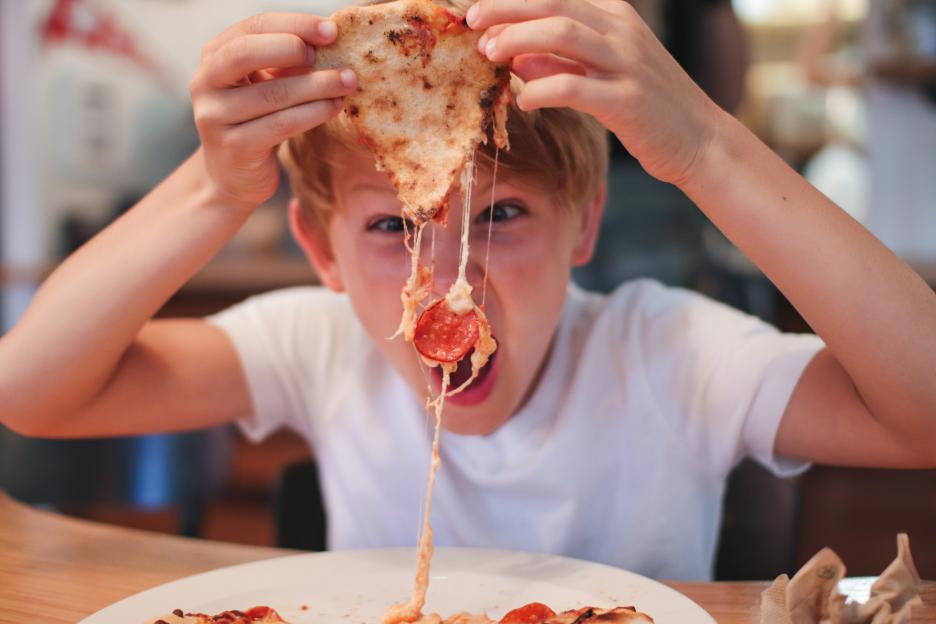 A young boy in a white t-shirt eating a slice of pepperoni pizza with cheese stretching from his mouth.
