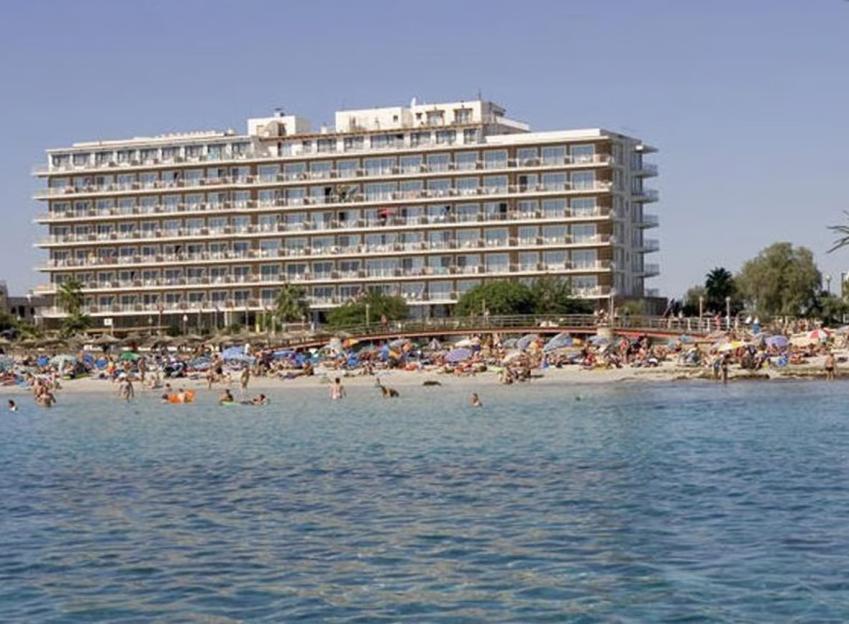 A large hotel overlooking a beach crowded with people and umbrellas, with a view of the clear blue sea.