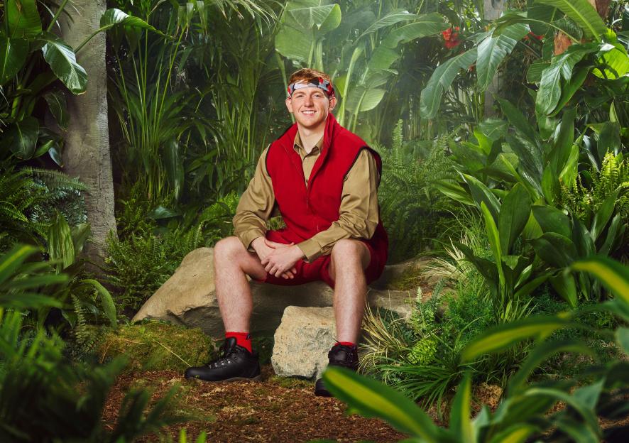 Angry Ginge from "I'm A Celebrity... Get Me Out Of Here!" sitting on a rock in a jungle setting.