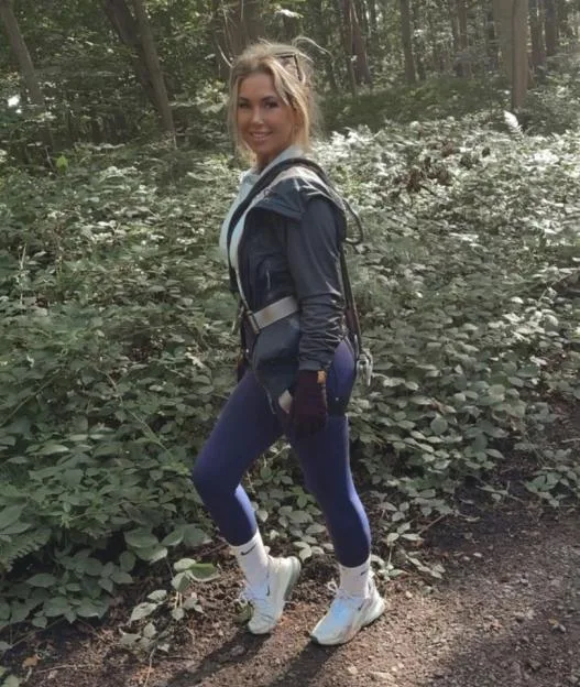 A woman with blonde hair in a ponytail wearing a black jacket, blue leggings, and white sneakers stands on a dirt path in a wooded area.