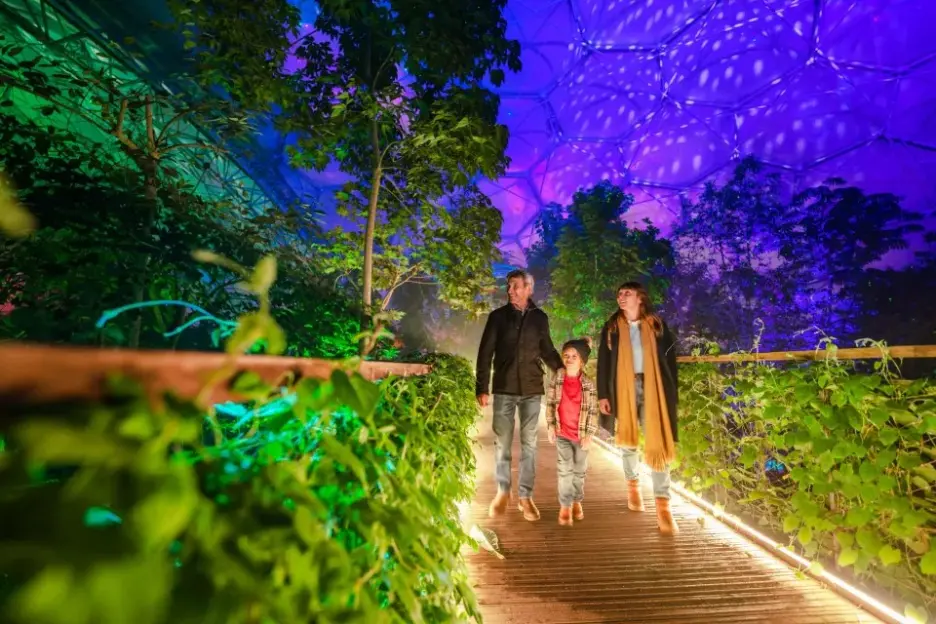 A family of three walks on an illuminated wooden path inside a large biodome with colorful lights on the plants and dome ceiling.