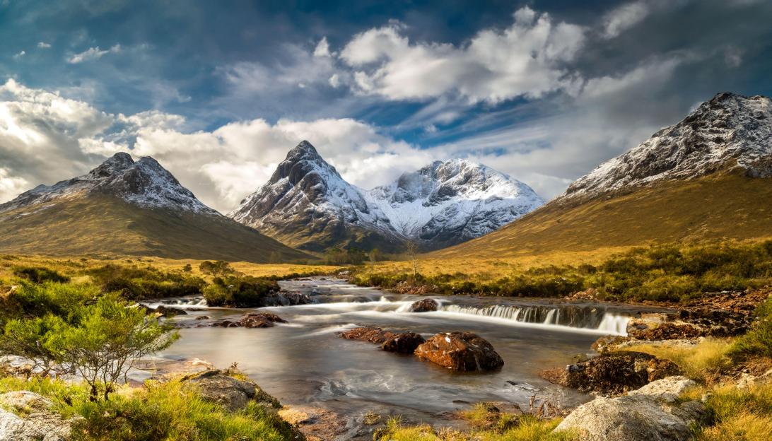 Long exposure photo of Buachaille Etive Mòr in Glencoe, Scotland, with a river flowing over rocks in the foreground and snow-capped mountains under a partly cloudy sky.