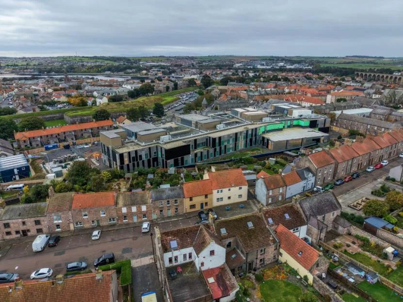 An aerial view shows the large, modern Berwick Community Hospital under construction, towering over the surrounding historic residential neighborhood in Berwick-upon-Tweed.