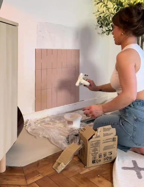 A woman applying mortar and rectangular tiles to a faux fireplace wall.