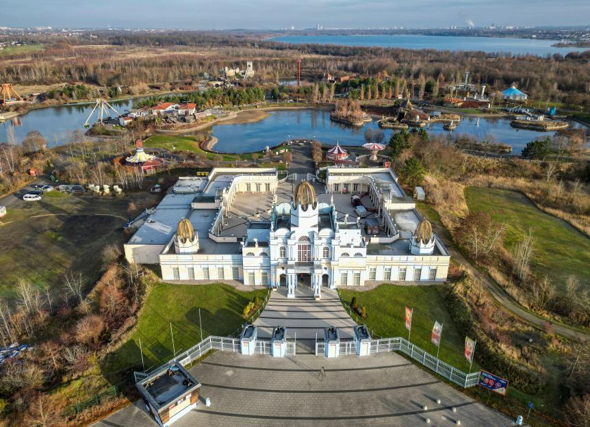 Aerial view of the entrance area of Belantis theme park, with a large, white classical-style building with golden domes, and various rides and a lake in the background.