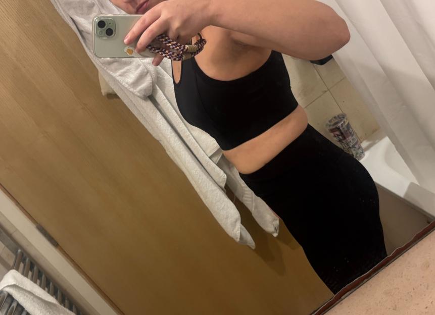 A person in a black bra and pants takes a mirror selfie to show their side profile.