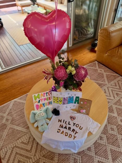 A white t-shirt with "WILL YOU MARRY DADDY" in gold letters, next to a pink heart balloon and flowers.
